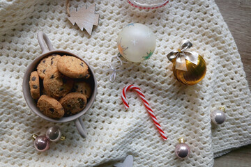 Bowl of cookies, soft knitted blanket and colorful Christmas ornaments at home. Flat lay.