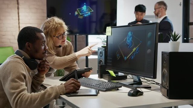 A European woman and an African American male artist discuss the 3D model in a computer game and application development studio among other team members.