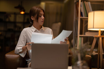 Asian woman sits at her desk with paperwork and computer at home working late at night.