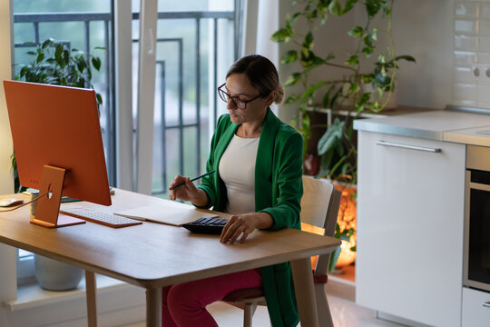 Focused woman counting profit on calculator near computer on workplace at home. Female freelancer counts expenses, analysing benefits counting budget, paying bills, taxes, rent, writes notes on paper.