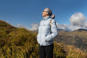 A gray-haired woman in sportswear and with a backpack stands high in the mountains against the background of clouds