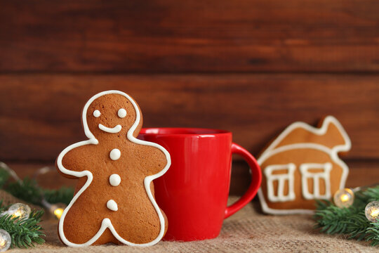 Christmas festive composition. Gingerbread man cookie and house coockie, red cup with tea or coffee, Christmas tree branches with lights against wooden background. Selective focus