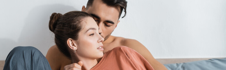shirtless man kissing girlfriend in t-shirt looking away in bedroom, banner.