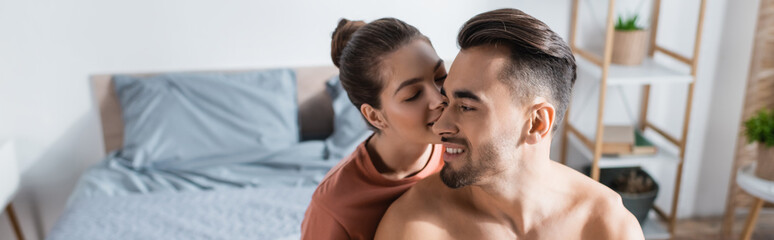 young woman kissing shirtless smiling man in blurred bedroom, banner.
