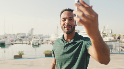 Happy young man making video call from phone while sitting in harbor with yachts on background