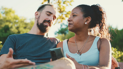 Happy interracial couple talking while sitting on bench, using cellphone