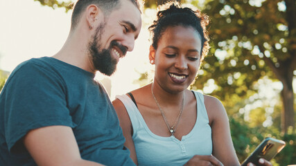 Happy interracial couple talking while sitting on bench, using cellphone