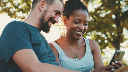 Happy interracial couple talking while sitting on bench, using cellphone