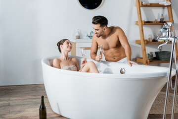 woman in bathtub and shirtless muscular man holding champagne glasses and smiling at each other.
