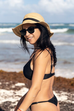 Beautiful and sexy Caucasian woman in a black bikini and a wicker hat walking along the seashore on a summer day.