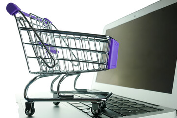 Laptop with small shopping cart on the white background, close up. A toy grocery cart on a laptop keyboard. The concept of electronic online purchases, e-commerce.