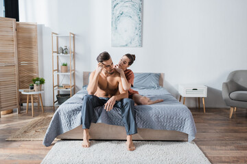 young woman embracing shirtless boyfriend sitting in pajama pants in spacious bedroom.