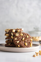 Cracked chocolate chip cookies with pieces of roasted peanuts on a ceramic plate with nuts in a blur on the background. Close-up with space for text.