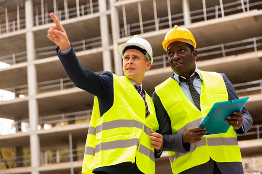 Young civil engineer discussing a construction plan with a colleague holding a folder of documents shows him something, pointing is it