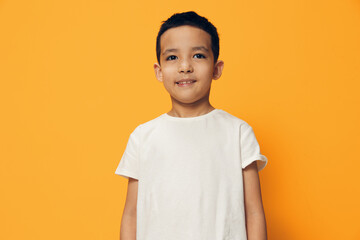 a cute, pleasant, smiling boy happily stands in a white cotton T-shirt on an orange background with an empty space for an advertising layout and looks pleasantly at the camera smiling.