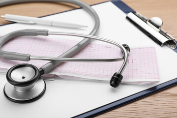 Clipboard with stethoscope and cardiogram on wooden table, closeup