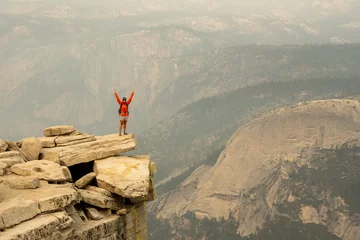 Papier Peint photo Half Dome Woman In Orange Coat Stands With Arms Raised On Half Dome Ledge
