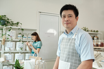 Fototapeta premium Portrait of an Asian male shopkeeper smiling and looking at camera, arranges natural products at refill store, zero-waste grocery, and plastic-free, eco environment-friendly, sustainable lifestyles.