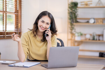 Freelance Smiling indian female young asian business woman using computer laptop and Talking Phone Working on Laptop In home Office.