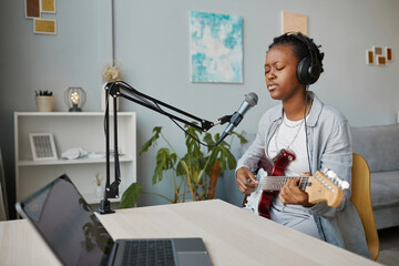 Minimal portrait of young black woman singing to microphone and playing guitar at home recording...