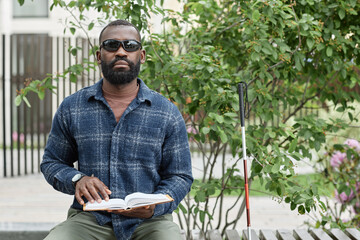 Fototapeta premium Front view portrait of blind man wearing sunglasses and reading book in braille while sitting on bench in park, copy space