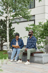 Vertical full length portrait of adult couple with partner with visual disability sitting on bench...