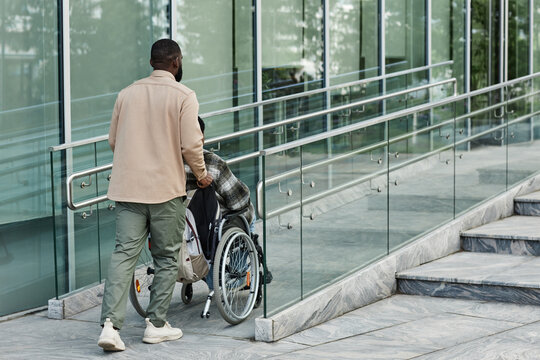 Back view of adult man assisting partner in wheelchair going up accessibility ramp in city, copy space