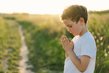 Boy closed her eyes and praying in a field at sunset. Hands folded in prayer concept for faith,...