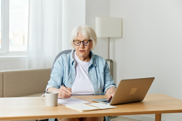 Fototapeta na wymiar a happy, successful elderly woman is sitting at her desk at home, stylishly dressed in black glasses and happily looking into the camera with a laptop and writing papers on the table
