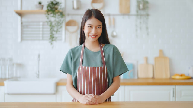 Happy young asian woman wearing apron and standing in kitchen room. Beautiful female smiling and looking at camera feeling confident
