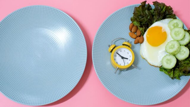 Female hand puts an alarm clock on plate of food on a pink background, concept of intermittent fasting.