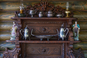 On a carved shelf stands silverware. In the old cupboard there is a teapot, a coffee pot, a sugar bowl, a tray, a vase, a saucepan, antique kerosene lamps and figurines.
