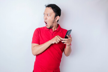 Surprised Asian man wearing red t-shirt pointing at his smartphone, isolated by white background