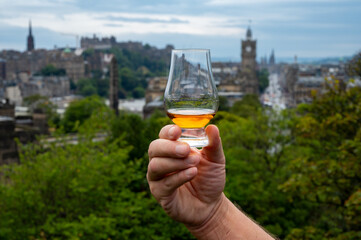 Hand holding glass of single malt scotch whisky and view from Calton hill to park and old parts of Edinburgh city in rainy day, Scotland, UK