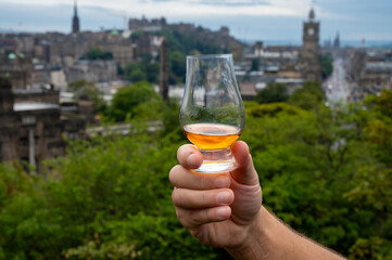 Hand holding glass of single malt scotch whisky and view from Calton hill to park and old parts of Edinburgh city in rainy day, Scotland, UK