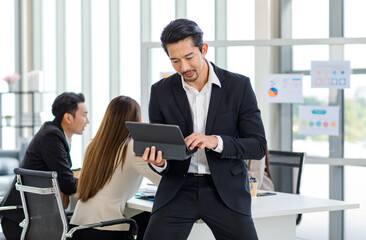 Millennial Asian professional successful bearded male businessman in formal suit sit on table browsing surfing internet via tablet computer in company office meeting room while colleagues discussing