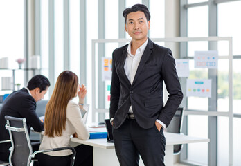 Millennial Asian professional successful young handsome businessman in formal suit standing posing put hands in pants pockets while male female colleagues discussing in company office meeting room