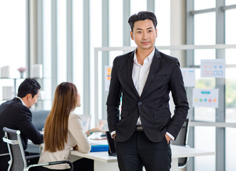 Millennial Asian professional successful young handsome businessman in formal suit standing posing put hands in pants pockets while male female colleagues discussing in company office meeting room