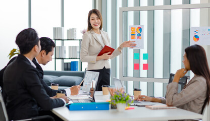 Millennial Asian professional successful young cheerful female  businesswoman speaker lecturer in formal suit standing smiling pointing graph chart information document on glass board in meeting room