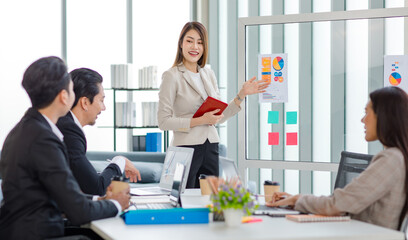 Millennial Asian professional successful young cheerful female  businesswoman speaker lecturer in formal suit standing smiling pointing graph chart information document on glass board in meeting room