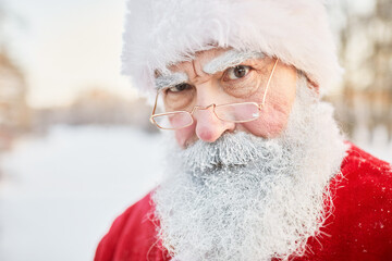Closeup portrait of serious Santa Claus looking at camera over eyeglasses in winter setting outdoors, copy space