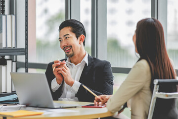 Millennial Asian professional successful bearded male businessman in formal suit sitting helping supporting discussing female colleague working with laptop notebook computer in company workstation