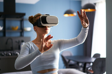 Woman in virtual reality goggles enters metaverse, controls immersive experience with hand gestures via headset interface in minimalistic interior. Girl gaming in cyber space. Futuristic concept.