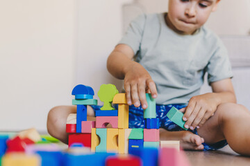 Happy caucasian boy sitting on floor, building tower from wood bricks and blocks. Playtime concept. Playroom, having fun on weekend, family activity.