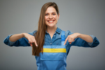 Smiling woman in blue overalls pointing finger down. Isolated female portrait in builder worker uniform.