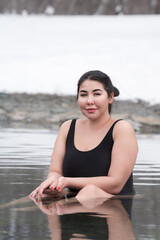 Fototapeta na wymiar Busty curvy plus size young adult model in black one-piece bathing suit sitting in geothermal water in outdoors pool at balneotherapy health spa, hot springs resort having balneological properties