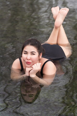 Overweight young woman in black one-piece bathing suit lying down, relaxation in geothermal mineral water in outdoors pool balneotherapy health spa, hot springs resort having balneological properties - obrazy, fototapety, plakaty