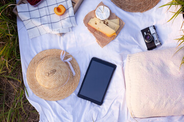 Fototapeta na wymiar Mockup of tablet lying on a picnic blanket with fruits, cheese, camera and straw hat, top view. Concept of leisure, technology and summer. Copy space. Gadgets on vacation. Remote work at summertime.