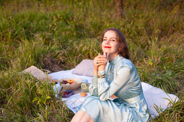 A girl in a long summer dress with short hair sitting on a white blanket with fruits and pastries and enjoying the moment. Concept of having picnic in a city park during summer holidays or weekends. 