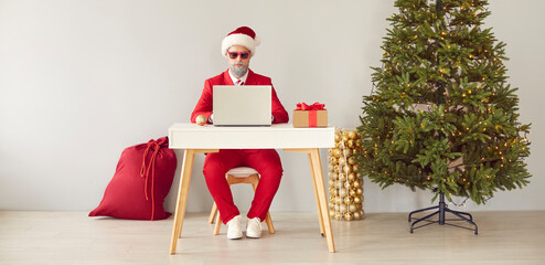 Funny man in red suit, Santa cap and sunglasses sitting at desk with Christmas present box in white...